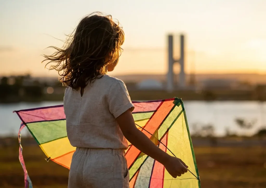 Criança de 3/4 de costas segurando pipa com horizonte de Brasília ao entardecer.