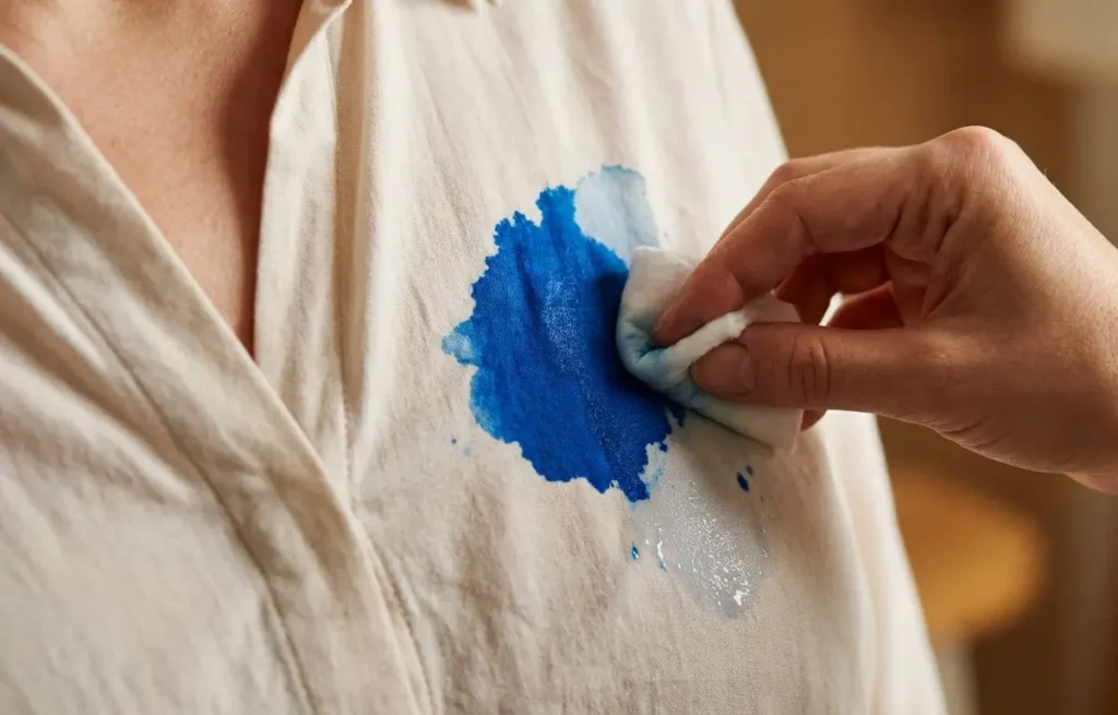 Mão aplicando removedor em mancha de tinta azul numa blusa clara, close-up.