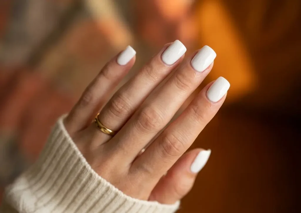 Mão com unhas de gel branca minimalista em fundo desfocado, anel dourado e manga creme
