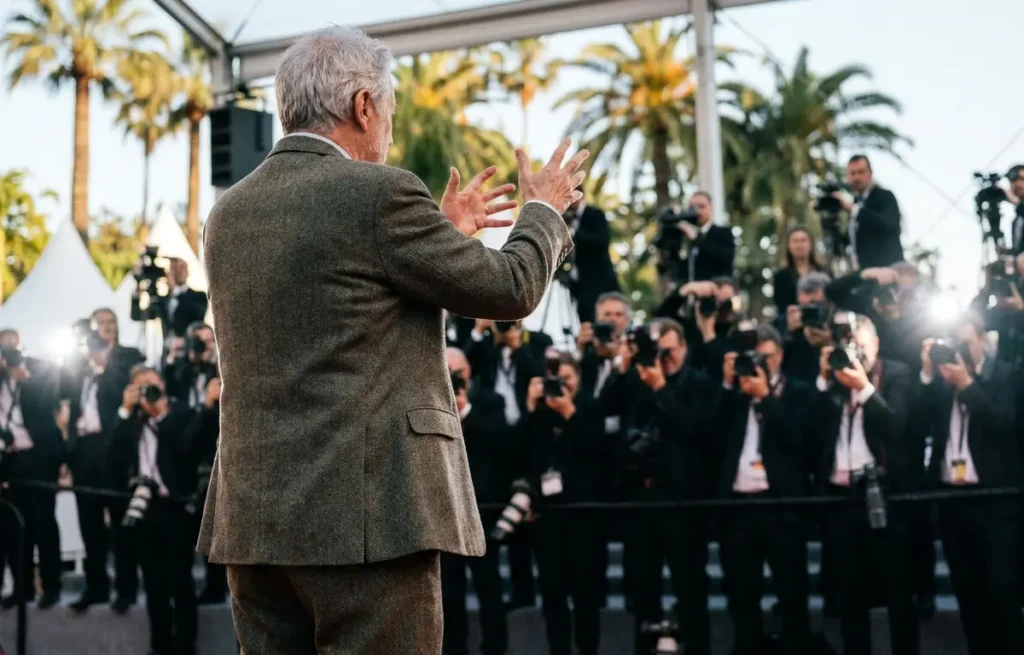 Cineasta em Cannes fotografado de costas em gesto enfático no tapete vermelho, fotógrafos desfocados ao fundo.