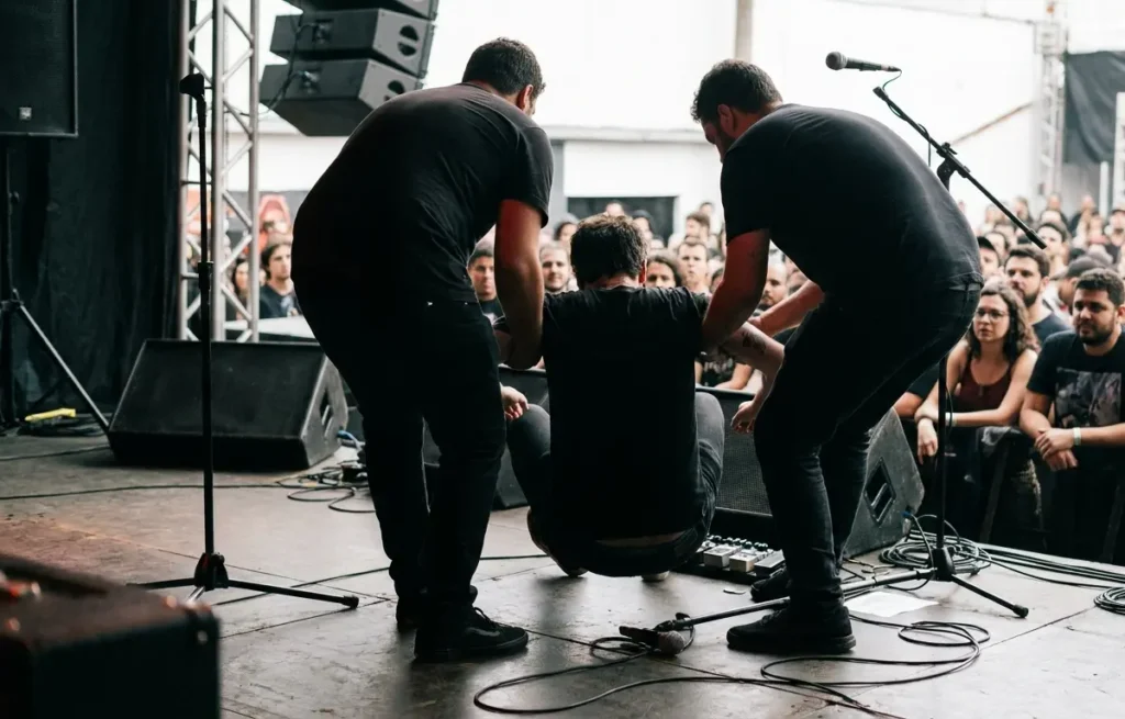 Músico sendo amparado no palco após queda visto de costas, com microfone caído