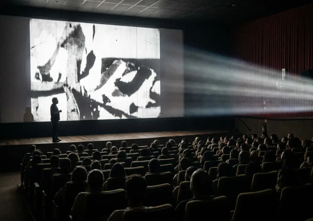Sala de exibição com público de costas, projetor iluminando a tela e silhueta do diretor em perfil.